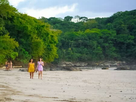 Zwei Frauen in bunten Kleidern gehen auf einem Sandstrand in Costa Rica auf die Kamera zu. Hinter ihnen kann man den Dschungel erkennen, sowie schwere Regenwolken, die über der Szene hängen.