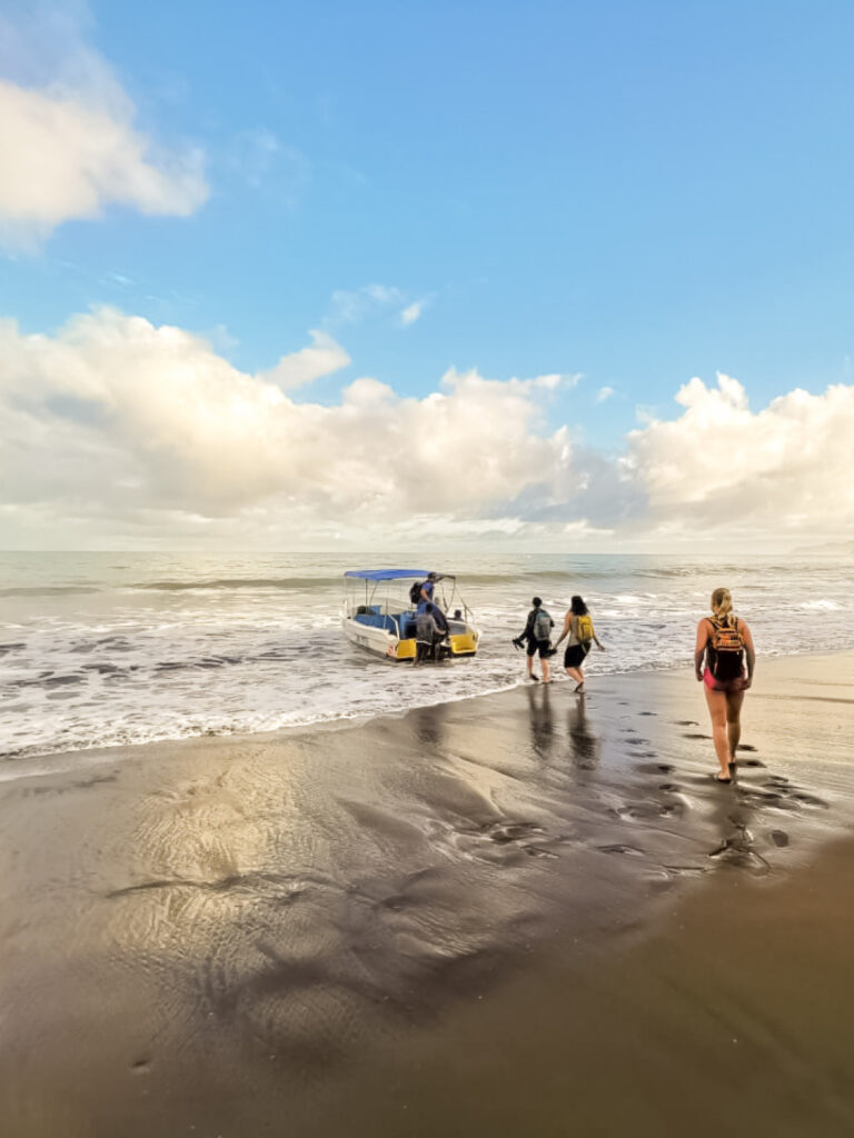 Mehrere Personen auf Wanderreise gehen auf einem Sandstrand im Nationalpark der Drake Bay auf ein kleines Boot zu, das auf den Wellen treibt. Die Sonne scheint vom Himmel herab.