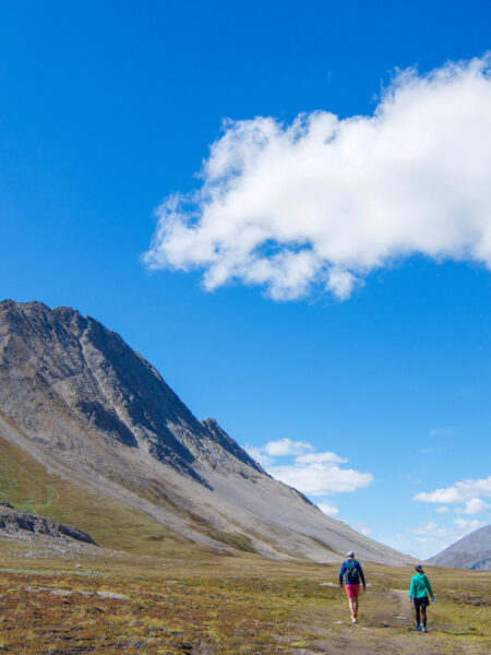 Zwei Personen auf einer Wanderreise durch den Westen von Kanada vor einem blauen Himmel mit einem Bergmassiv zu ihrer linken.