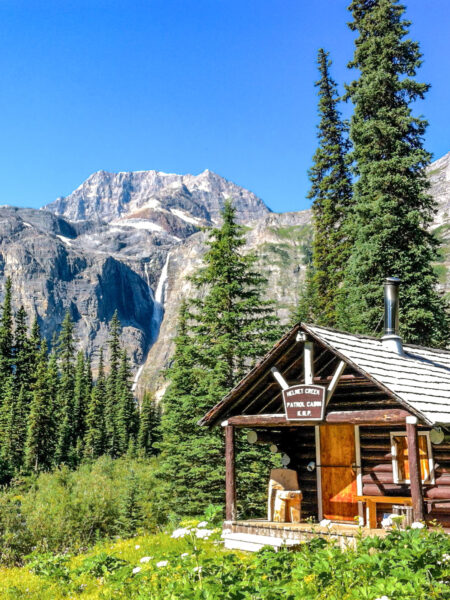 Eine Blockhütte in einem kanadischen Wald vor einem größeren Bergmassiv im Hintergrund.