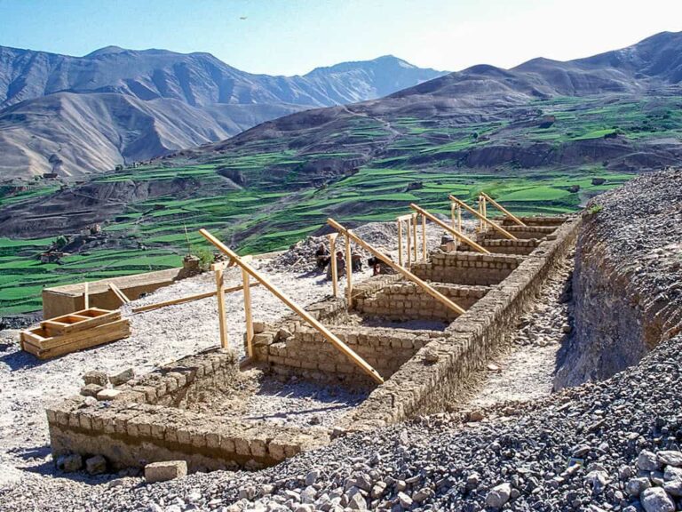 Steinfundamente eines Schulgebäudes in einer weiten Hochgebirgslandschaft in Ladakh, umgeben von grünen Feldern und kargen Bergen.