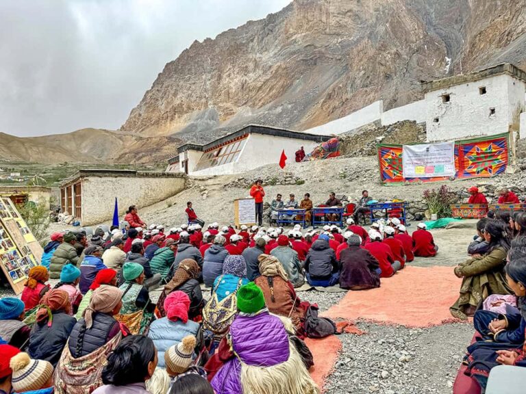 Viele Menschen sitzen bei einer Veranstaltung vor traditionellen Gebäuden in einem abgelegenen Bergdorf in Ladakh, eingerahmt von steilen Felswänden