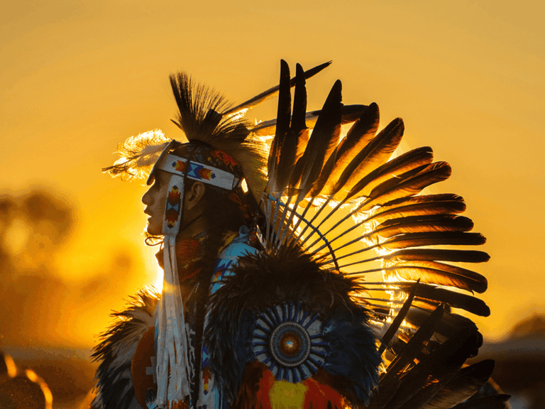 Indigener Tänzer in traditioneller Kleidung mit Federkopfschmuck im warmen Licht des Sonnenuntergangs.