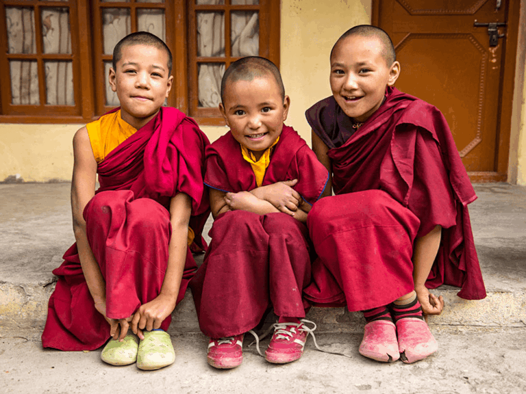 Drei junge buddhistische Mönche in roten Gewändern sitzen lächelnd vor einem Kloster in Ladakh.